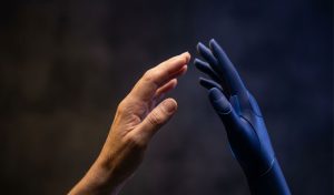 Une main humaine et une main artificielle face à face sans se toucher, dans une lumière contrastée aux tons indigo — le dialogue entre l'humain et l'IA.